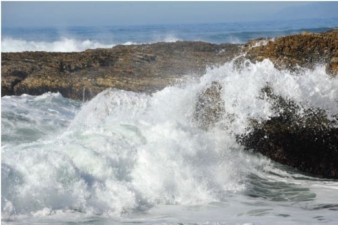 Below is a picture of water striking rocks at a coastline.    Describe which of Earth's  spheres  are interacting here, and some of the effects that may be caused by this intera