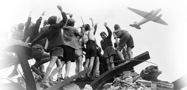     -What ironic element regarding the Berlin airlift of 1948-1949 does this photograph capture? A)  German children mistrusted Allied planes despite the fact that they were receiving food aid. B)  The food dropped on Berlin was limited to the same gruel that German children were already eating. C)  German children still used the Nazi salute to greet others despite Hitler's defeat. D)  German children cheerfully greet an American plane from the rubble that similar planes created just a few years before.