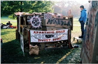 The image  Antiapartheid Protest, Cornell University  from 1986 shows students with a self-made shantytown hut resembling those in South Africa. What does this image suggest?   A)  American businesses employed South African workers under unspeakable conditions. B)  American businesses were involved in South African businesses. C)  Cornell students had brought their own experiences in South Africa home. D)  Cornell University was using South Africans for unethical research and experiments.